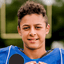A young male athlete wearing a blue football jersey, holding a black microphone in front of his face, with a blurred green outdoor background.