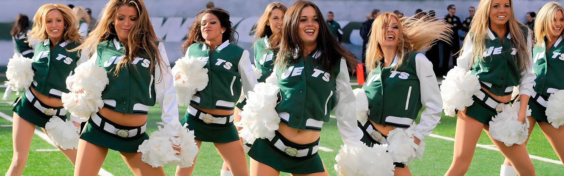 A group of cheerleaders in matching green and white jackets perform on a football field, holding white pom-poms.