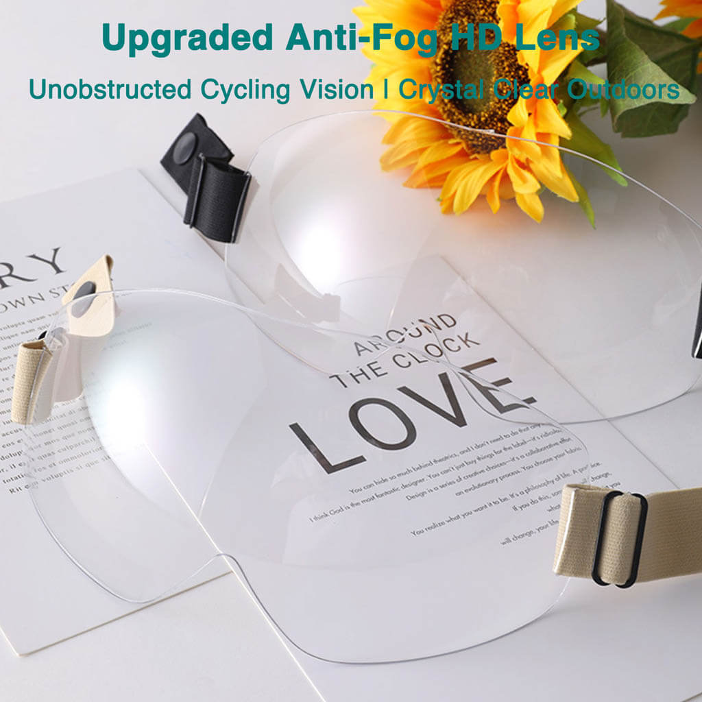 Two transparent anti-fog HD cycling lenses with adjustable straps are displayed on a white surface next to papers and a sunflower.