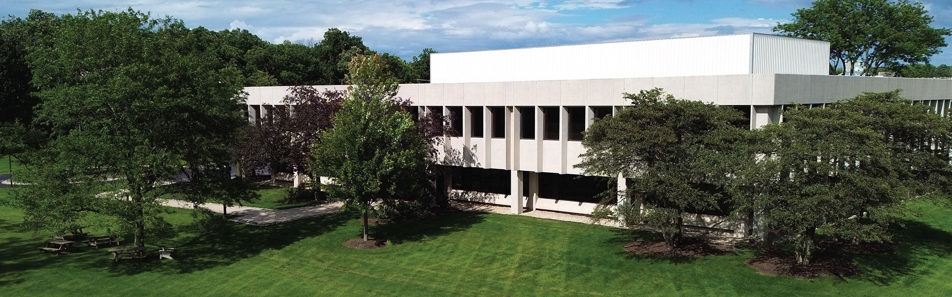A modern, two-story building with large windows and columns, surrounded by green lawns, mature trees, and picnic benches on a sunny day.