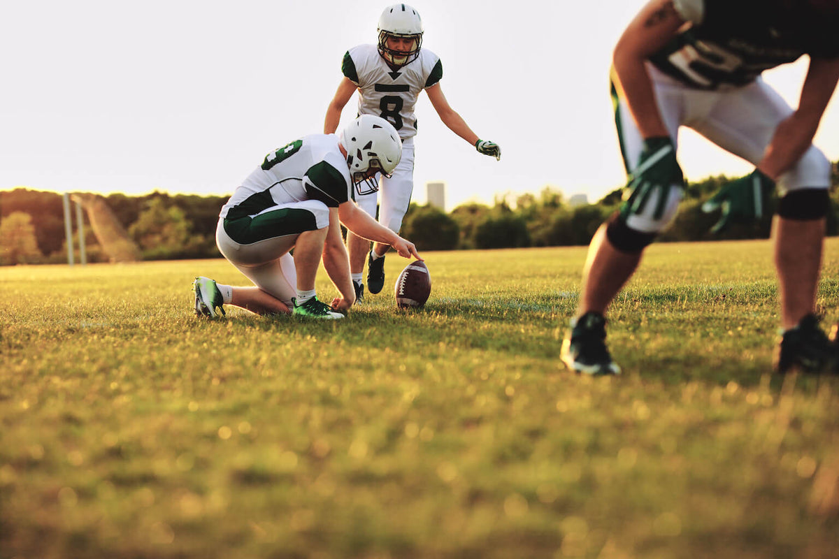 Three American football players on a field, with one holding the ball steady for a kick while another prepares to kick it. The sun is setting in the background.