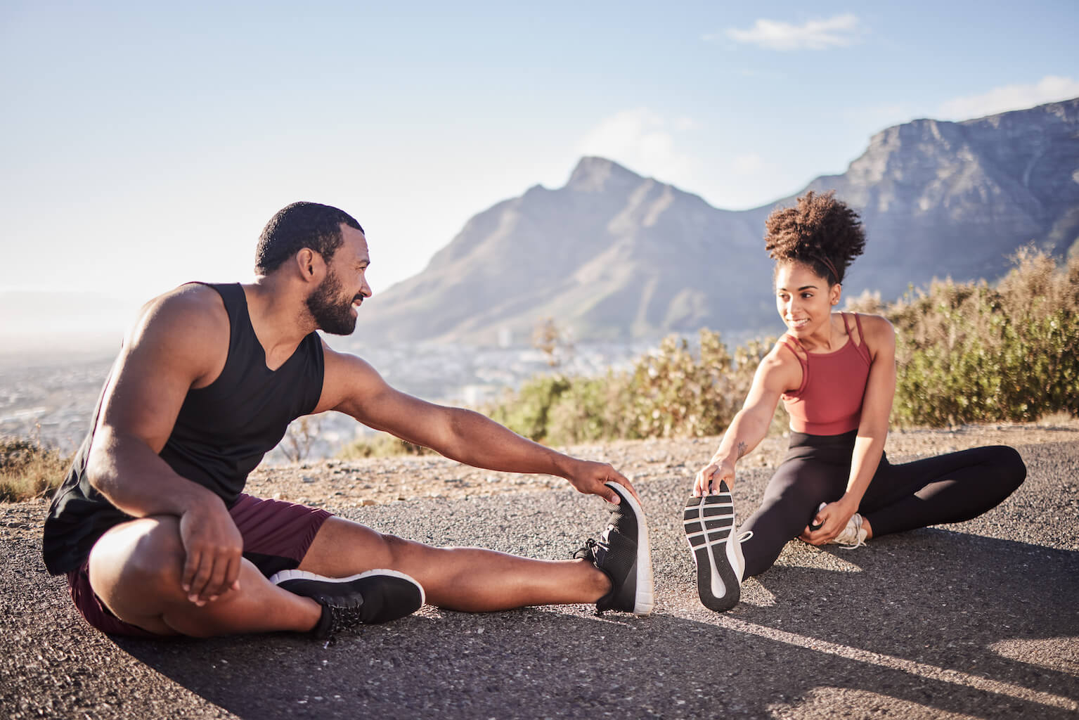 A man and woman stretching on the side of a road with mountains in the background.