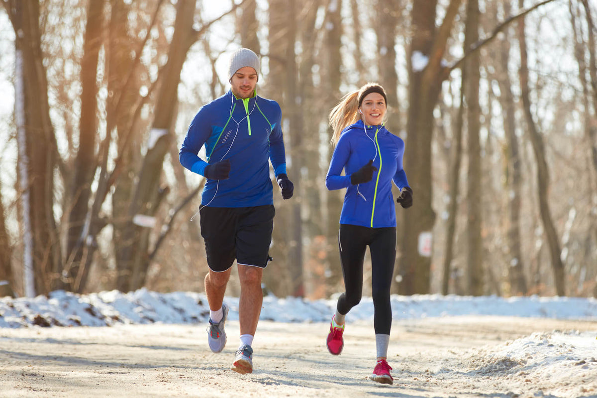 A man and a woman are jogging on a snow-lined path in a wooded area, wearing blue athletic gear and gloves.