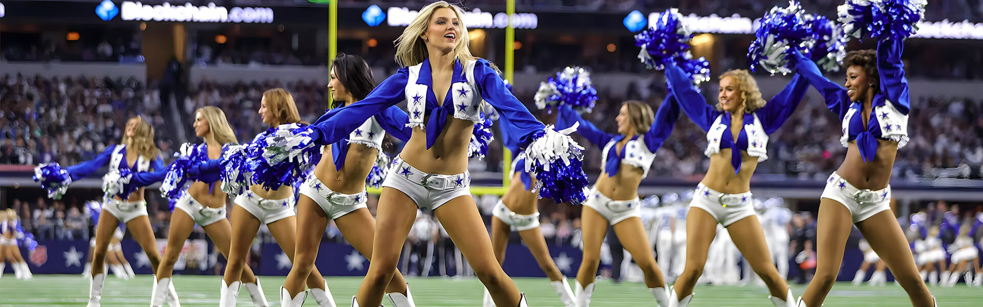 Cheerleaders in blue and white uniforms perform a dance routine on a football field, pom poms in hand and a stadium crowd cheering enthusiastically in the background.