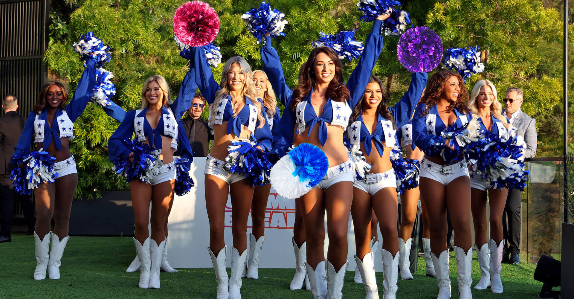 A group of cheerleaders in blue and white uniforms perform outdoors, raising blue and white pom poms in the air.