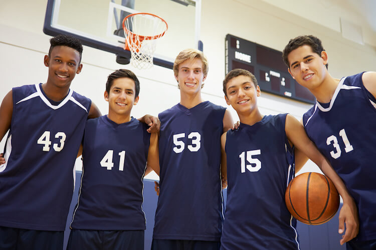 Five male basketball players standing side by side with hands on each other’s shoulders and smiling