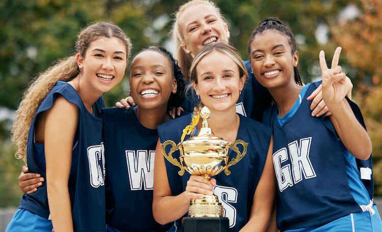 Five female players in blue jerseys holding a trophy and smiling