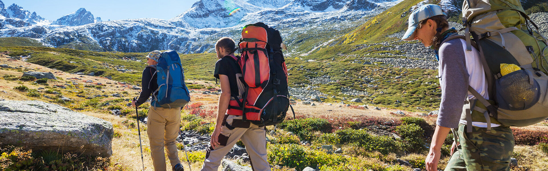 Three hikers with backpacks walk uphill on a sunny day in a mountainous landscape with snow-capped peaks in the background.